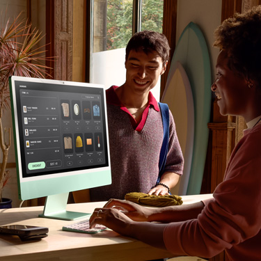 Pracownik siedzący za ladą w sklepie z deskami surfingowymi i innymi akcesoriami finalizuje na iMacu zakup dokonany przez klienta stojącego po drugiej stronie lady.