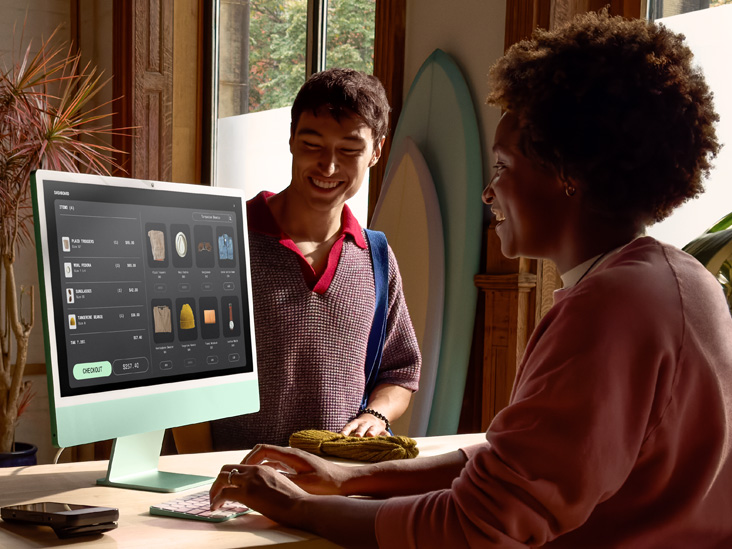 Two people in a well-lit room, one checking out an order on a green iMac M4.