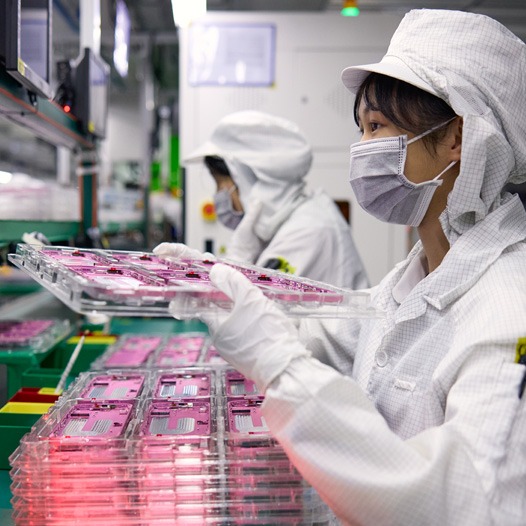 Two employees wearing full-coverage work suits handle iPhone parts in an assembly facility